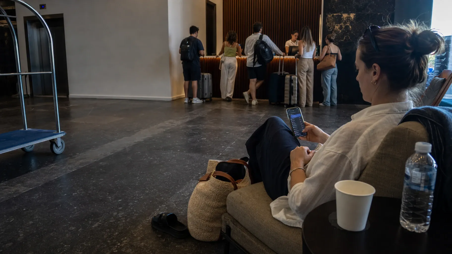 A guest relaxes in a lobby armchair using her phone while other guests queue with suitcases at the reception desk behind her
