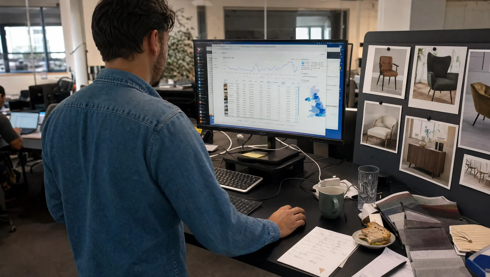A marketing manager at a standing desk studies a campaign dashboard with spend by day and a regional map, photos of chairs pinned beside him