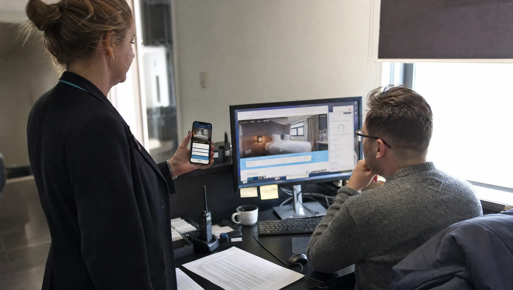 Two hotel staff review a new website before launch: the mobile version on her phone, the room page and a speed report on his monitor